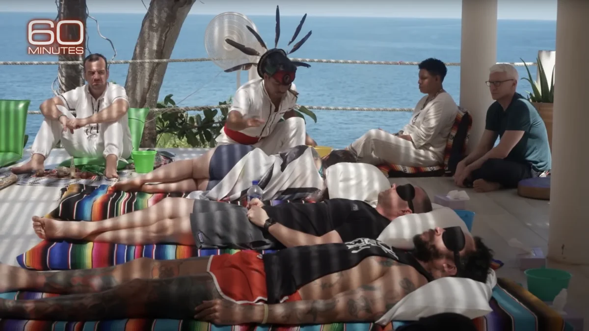 A group relaxes on colorful blankets by the ocean, while a healer performs a ritual nearby.